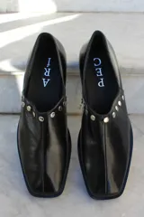Zapato de cuero vacuno color bordó, con diseño de punta cuadrada y detalle de tachas metálicas planas en el borde superior. Presenta un taco geométrico bajo y costura central en el empeine.