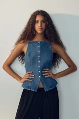 Chaleco sastrero en denim de algodón rígido, con escote recto, breteles finos y cierre frontal de botones metálicos. Presenta costuras princesa al frente y una abertura pronunciada en el dobladillo inferior.
