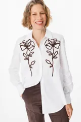 Camisa blanca de corte clásico con cuello camisero y cierre frontal de botones. Presenta un bordado lineal en color marrón oscuro en ambos lados del frente, representando motivos florales.
