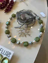 Collar artesanal compuesto por cuentas cuadradas de color verde oliva, cuentas de jaspe dálmata, perlas de río irregulares y un dije central metálico con forma de sol. Incluye un cordón de gamuza gris ajustable y terminaciones en acero.
