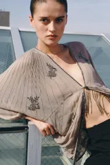 Top de viscosa color beige con escote en V, mangas murciélago abullonadas por debajo del codo y bajo asimétrico. Presenta delicados bordados con aplicaciones de abalorios y cuentas brillantes en el frente y las mangas.