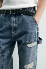 Pantalón de jean estilo carpintero con corte relaxed fit, tiro medio y pierna recta. Presenta un lavado azul medio con efecto desgastado y roturas en las rodillas. Incluye bolsillos laterales tipo plastrón, tira multifunción en la pierna, cierre frontal con cremallera y botón.