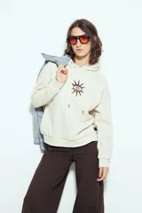 Canguro color crema de calce relajado, con capucha y cordones ajustables. Presenta un estampado gráfico frontal de un ojo con rayos.