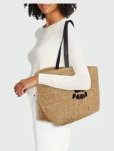 Tote bag de rafia natural con diseño tejido, asas largas de cuero negro y bordado frontal con el logo de la marca en hilo negro.