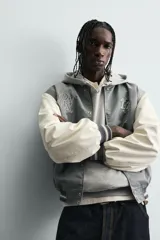 Cazadora estilo varsity o bomber, cuerpo principal gris oscuro y mangas color crema. Presenta cuello, puños y bajo en rib elástico con rayas grises y blancas. Tiene bordados en el pecho: un monograma estilizado a la izquierda y el texto "EST. 1975" a la derecha.