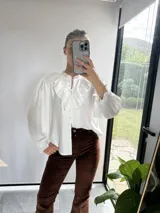 Camisa blanca de tela liviana con cuello mao y volados frontales de encaje bordado. Presenta mangas largas abullonadas con puños elásticos y cierre frontal con botones.