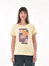 Remera de manga corta color amarillo pálido, con cuello redondo y un estampado frontal que incluye flores estilizadas en tonos naranja, morado, rosa y negro, con el texto "FLOWER FIELDS".