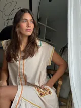 Vestido corto beige de algodón con cuello camisero y cierre frontal de botones. Presenta mangas cortas y detalles bordados a contratono en la zona del pecho y en los bolsillos, además de un volado en el ruedo.