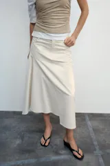 Falda midi de tiro medio color gris, con diseño asimétrico en el bajo, bolsillos frontales y traseros, y cierre frontal con cremallera y botón.