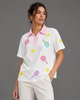 Chomba oversize blanca con cuello polo en color rosado. Presenta un estampado integral de raquetas de tenis y pelotas en colores vibrantes como amarillo, fucsia, turquesa y violeta.