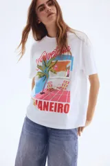 Remera blanca de algodón con cuello redondo y hombros caídos, con un estampado gráfico que muestra una escena de playa o terraza con una palmera, sillas de lona a cuadros y el texto "Sorveteria Rio de Janeiro".
