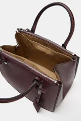 Bolso pequeño tipo satchel, color borgoña oscuro, con doble asa de mano y correa bandolera extraíble. Presenta dos compartimentos exteriores con cierre de cremallera metálica y un compartimento interior con cierre imantado.