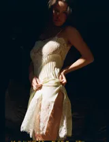 Vestido midi de estilo lencero confeccionado en viscosa satinada color crema. Presenta escote recto con tirantes finos, detalle de encaje floral en el pecho y el ruedo, y pliegues verticales en el frente.