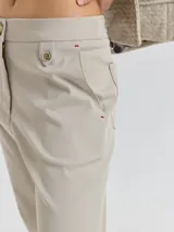 Pantalon de corte cigarette color beige, con cierre frontal, bolsillos laterales y detalle de botón decorativo en la cintura. Presenta costuras reforzadas con hilo rojo en los extremos de los bolsillos.