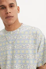 Remera de manga corta con cuello redondo y calce estándar. Presenta un estampado geométrico con líneas horizontales en tonos pasteles, azul y amarillo.