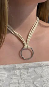 Gargantilla tipo choker compuesta por múltiples tiras finas de gamuza en color crema, que se unen en el centro a través de una argolla metálica plateada.