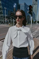 Camisa de popelín blanca con cuello solapa decorado con puntilla y lazo frontal. Presenta mangas largas con puños abotonados y cierre frontal con botones.