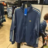 Campera deportiva Adidas Originals de color azul, con cierre frontal, cuello alto y bolsillos laterales. Presenta las icónicas tres tiras en los hombros y mangas en tonos azul claro y blanco, junto con el logo del trébol bordado en dorado en el pecho.