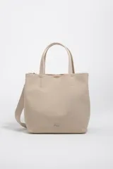 Bolso de mano grande tipo tote de Bimba y Lola, color beige, con doble asa superior y correa ajustable de tejido. Presenta logo metálico en la parte inferior y cierre superior.