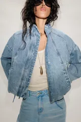 Cazadora bomber de denim con cuello subido, manga larga y cierre frontal con cremallera y botones a presión ocultos. Presenta costuras verticales decorativas y bajo con ajuste elástico.