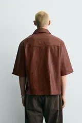 Sobrecamisa de corte relajado confeccionada en tejido efecto piel, con cuello solapa, manga corta, bolsillo plastrón en el pecho y cierre frontal con cremallera.