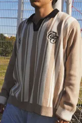 Buzo de punto tejido en jacquard con diseño de rayas verticales en tonos beige y blanco. Presenta cuello polo en color negro, puños y ruedo acanalados en blanco, y bordados alusivos al mundial en el pecho.