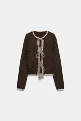 Chaqueta de punto de textura bouclé en color marrón oscuro, con cuello redondo y manga larga. Presenta detalles de vivo a contraste en color crema en los bordes y cierre frontal mediante lazadas decorativas con flecos.