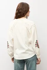 Camisa de color natural con mangas largas abullonadas y bordado floral multicolor en los puños. Presenta cuello mao y cierre frontal con botones.