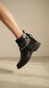 Bota corta de cuero negro con taco cuadrado bajo. Presenta un diseño versátil con un accesorio desmontable que incluye flecos de cuero, tachas metálicas y una cadena decorativa alrededor del tobillo.