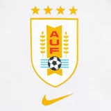Remera blanca de algodón con cuello redondo y manga corta. Presenta un escudo estilizado de la Asociación Uruguaya de Fútbol (AUF) con cuatro estrellas doradas arriba y el logo de Nike en color dorado.