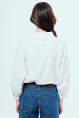 Camisa blanca de manga larga abullonada con puños elásticos, cuello camisero clásico y cierre frontal de botones. Presenta detalles de alforzas y encaje geométrico en la pechera.