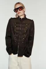 Chaqueta de gamuza marrón con cuello alto, manga larga y bajo con peplum. Cuenta con doble botonadura decorativa de botones metálicos dorados en el frente y cierre con ganchos ocultos.