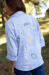 Camisa de lino color celeste, con cuello clásico, cierre frontal de botones y detalle de cuatro estrellas bordadas en color amarillo sobre el pecho. Presenta mangas tres cuartos con detalle de encaje en los puños.