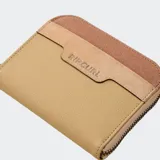 Billetera compacta con cierre de cremallera, color beige y marrón. Presenta paneles de diferentes texturas y el logo de Rip Curl grabado en el frente.