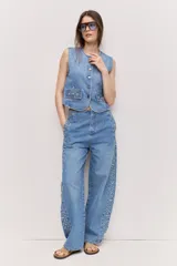 Chaleco de denim azul sin mangas, con escote redondo, cierre frontal de botones metálicos y bolsillos delanteros decorados con tachas.
