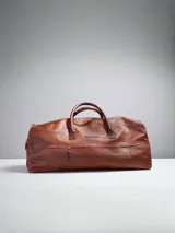 Bolso de viaje tipo weekender de cuero color marrón, con dos asas superiores y un bolsillo exterior con cierre en el frente. Presenta un parche de marca cosido en el centro superior.