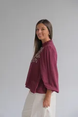 Blusa color bordó con bordados florales en tono crema en la parte superior frontal. Presenta cuello redondo con pequeña abertura, cierre frontal con botones y mangas largas acampanadas con puños ajustados.