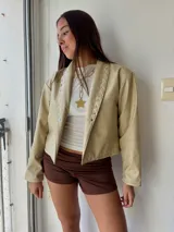 Campera de ecocuero color crudo, con diseño de solapas decoradas con tachas metálicas circulares a lo largo de los bordes frontales. Presenta un corte recto y mangas largas.