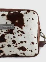 Morral de cuero marca Tranquera con frente de pelo natural en diseño animal print de vaca. Presenta una estructura rectangular con bordes redondeados, correa de cuero regulable y desmontable mediante mosquetones metálicos, y una placa con el logo de la marca en el centro.