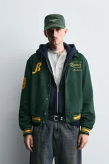 Cazadora tipo varsity de corte relajado confeccionada en tejido efecto ante color verde oscuro. Presenta cuello solapa, cierre frontal con botones a presión y puños y bajo acanalados con rayas en contraste. Incluye parches bordados en color amarillo en el frente y mangas.
