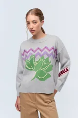 Sweater gris de tejido de punto con cuello redondo y mangas largas. Presenta bordados de una flor verde y la palabra "Rebels" en la manga.