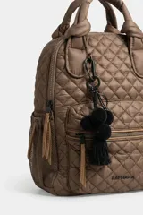 Mochila matelaseada de color marrón, con diseño acolchado de rombos, asas superiores con nudos y detalles en tono tostado. Cuenta con un bolsillo frontal con cierre, bolsillos laterales de red y un llavero decorativo con pompones y borla negra.