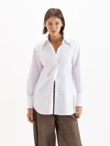 Camisa holgada de poplin blanco, con cuello clásico, mangas largas con puños anchos y cierre frontal con botones. Presenta detalles de apliques metálicos en las puntas del cuello y sutil bordado en el delantero.
