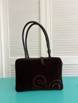 Bolso de mano o de hombro de forma rectangular, confeccionado en material tipo gamuza color marrón oscuro, con asas de cuero marrón oscuro. Presenta un diseño bordado o grabado en el frente con letras estilizadas en tono marrón claro.