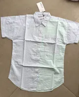 Camisa de diseño color-block a rayas, con un lado en tonos celeste y blanco y el otro en verde claro y blanco. Presenta mangas cortas, cuello camisero, cierre frontal con botones y un bolsillo aplicado en el pecho.