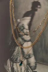Colgante dorado de tres cadenas de diferentes grosores y diseños. La cadena más larga tiene dos dijes circulares unidos. La cadena más corta tiene un dije de piedra facetada color verde oscuro.