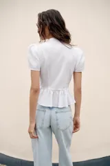 Camisa blanca de manga corta abullonada, con cuello mao y cierre frontal con alamares. Presenta corte evasé con detalle de volados en el bajo.