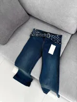 Pantalón jean de corte recto y tiro alto, color azul oscuro, con pretina ancha decorada con múltiples ojales metálicos y tachas, e incluye un cinturón incorporado con hebilla metálica.