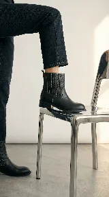Bota de caña alta color negro, con diseño versátil que incluye un arnés desmontable con hebilla y argollas metálicas. Presenta costuras decorativas en la parte superior de la caña y un taco bajo cuadrado.