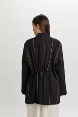 Chaqueta negra de corte oversize y diseño desestructurado, con solapas clásicas, cierre frontal de dos botones y sutil estampado de rayas verticales finas.